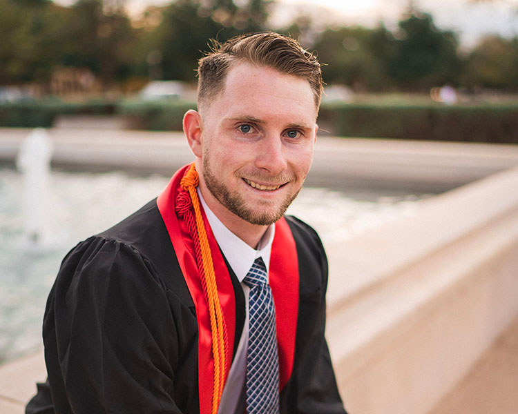 Male graduate sitting on a fountain and smiling.